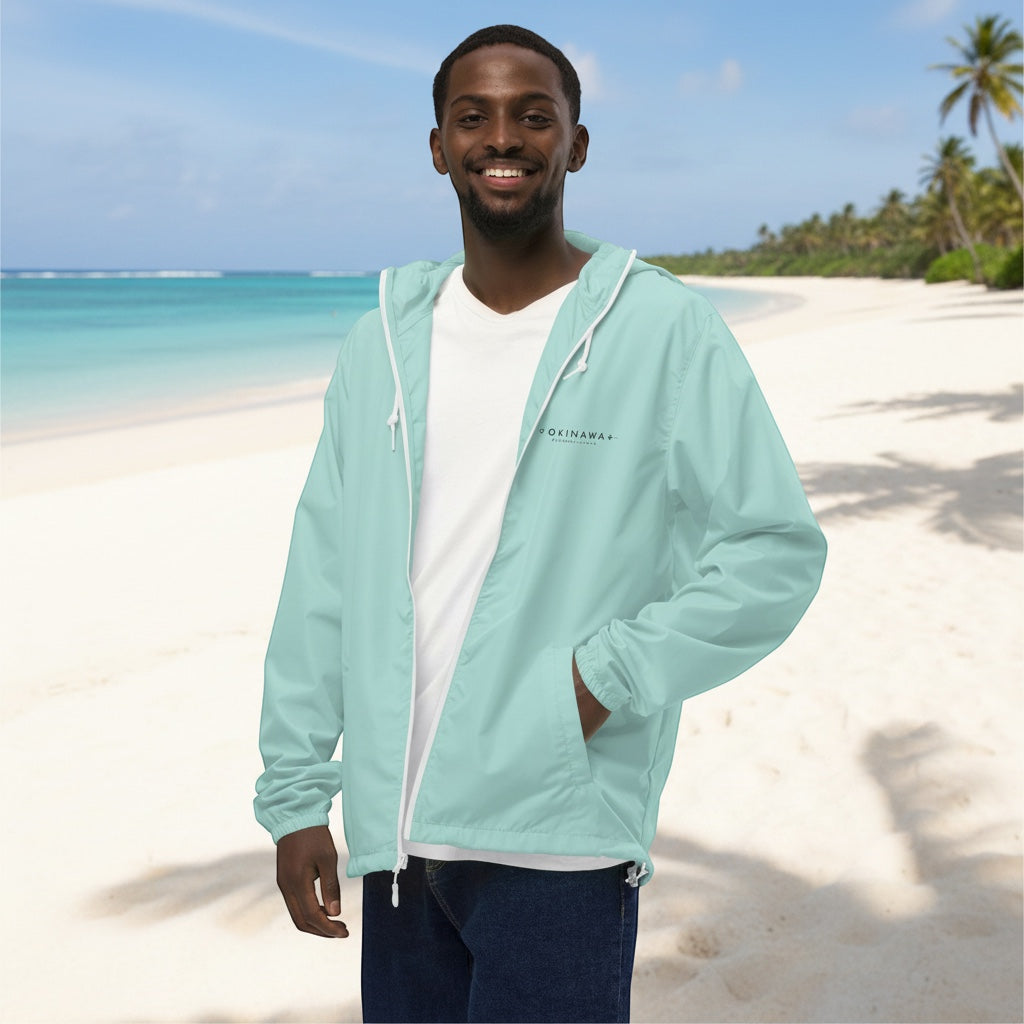 A man wearing a light blue YUTASHA Toshie windbreaker with a hood and white zipper on a sunny tropical beach.