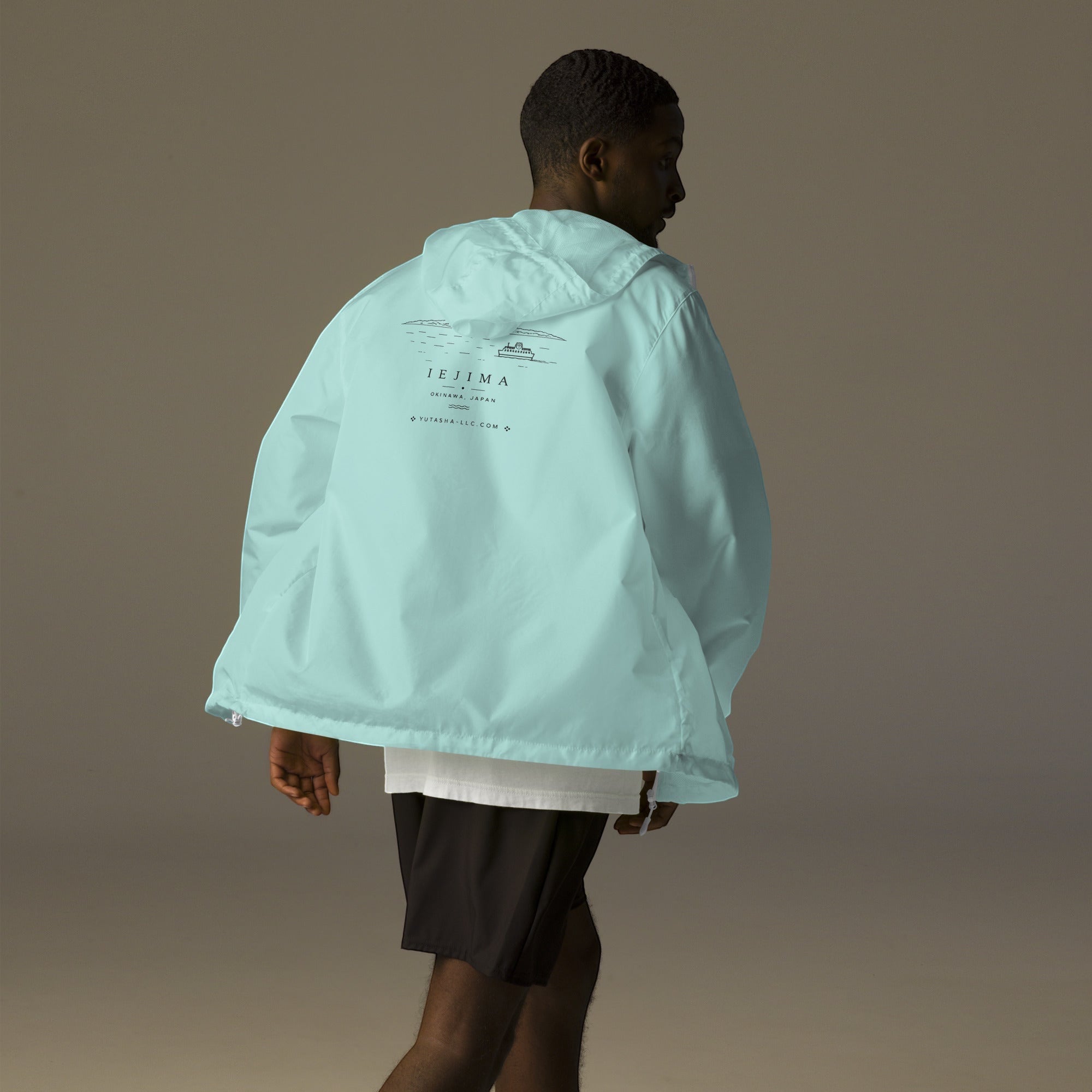 YUTASHA Toshie windbreaker in light blue with Iejima graphic print on back, worn by a man from behind.